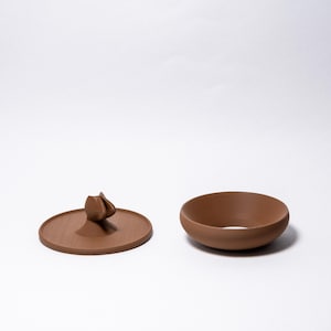 Peut inclure: Un bol en c&eacute;ramique marron et son couvercle assorti avec une poign&eacute;e d&eacute;corative. Le couvercle est rond et pr&eacute;sente un motif floral stylis&eacute; en relief. Le bol est de forme simple et arrondie.