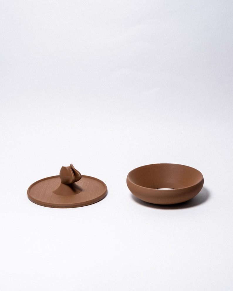 Peut inclure: Un bol en c&eacute;ramique marron et son couvercle assorti avec une poign&eacute;e d&eacute;corative. Le couvercle est rond et pr&eacute;sente un motif floral stylis&eacute; en relief. Le bol est de forme simple et arrondie.