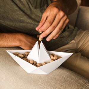 Puede incluir: Un cuenco blanco con forma de barco de papel lleno de pistachos. Se ve a una persona dejando caer pistachos en el cuenco. El cuenco está sobre una superficie beige. La persona lleva una camisa verde.