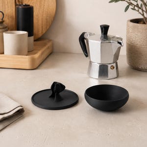 Peut inclure: Une cafeti&egrave;re italienne en argent avec une poign&eacute;e et un couvercle noirs est pos&eacute;e &agrave; c&ocirc;t&eacute; d'un bol noir et d'un couvercle noir avec une poign&eacute;e d&eacute;corative. Une planche &agrave; d&eacute;couper en bois et une petite plante en pot sont en arri&egrave;re-plan.