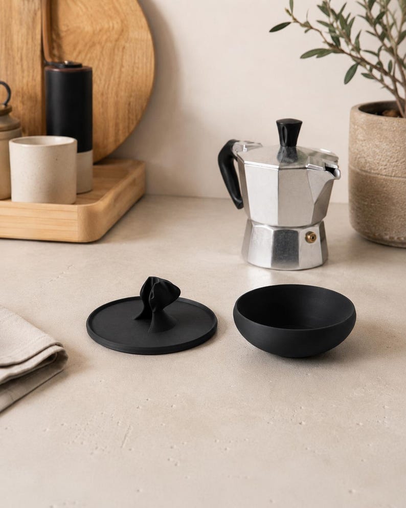 Peut inclure: Une cafeti&egrave;re italienne en argent avec une poign&eacute;e et un couvercle noirs est pos&eacute;e &agrave; c&ocirc;t&eacute; d'un bol noir et d'un couvercle noir avec une poign&eacute;e d&eacute;corative. Une planche &agrave; d&eacute;couper en bois et une petite plante en pot sont en arri&egrave;re-plan.