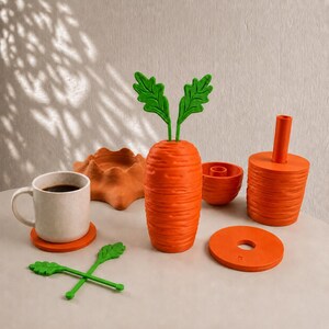 May include: A collection of orange and green carrot-themed items including a mug on a coaster, a textured carrot-shaped container with green leafy stems, a small bowl, a cylindrical holder, a flat circular disc, and two green leafy stirrers.