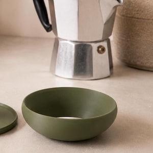 Peut inclure: Un bol en c&eacute;ramique vert olive avec un couvercle assorti. Une cafeti&egrave;re italienne argent&eacute;e est &agrave; l'arri&egrave;re-plan, ainsi qu'un r&eacute;cipient beige. Le bol a une finition lisse et mate et une forme arrondie, id&eacute;ale pour servir des aliments.