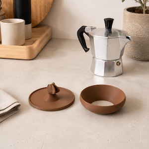 Peut inclure: Une cafeti&egrave;re italienne en argent avec une poign&eacute;e et un couvercle noirs, &agrave; c&ocirc;t&eacute; d'un couvercle en c&eacute;ramique marron et d'un bol en c&eacute;ramique marron. Les articles sont sur une surface claire.