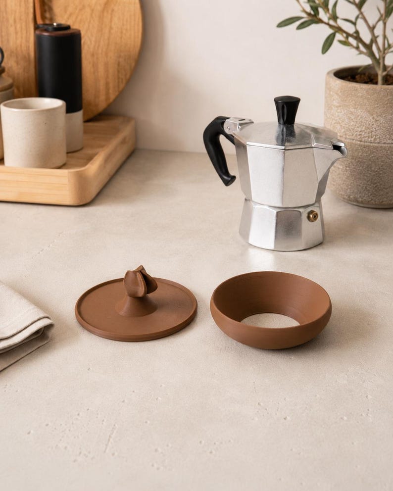 Peut inclure: Une cafeti&egrave;re italienne en argent avec une poign&eacute;e et un couvercle noirs, &agrave; c&ocirc;t&eacute; d'un couvercle en c&eacute;ramique marron et d'un bol en c&eacute;ramique marron. Les articles sont sur une surface claire.