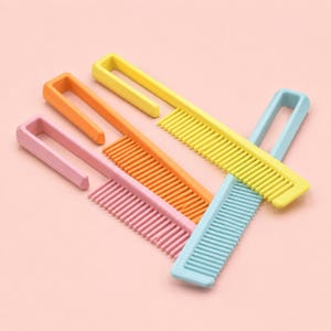 Peut inclure: Plusieurs peignes en plastique colorés, roses, oranges, jaunes et bleus. Chaque peigne a une poignée et des dents rapprochées. Les peignes sont disposés sur un fond rose clair.