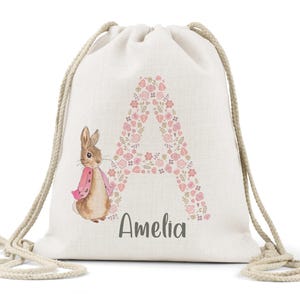 May include: A white drawstring bag with a pink floral letter A and a bunny rabbit illustration. The bag has the name Amelia printed below the letter.