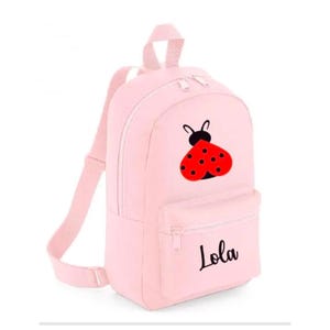 May include: A pale pink backpack featuring a ladybird design and the name "Lola" in black script. The ladybird is red with black spots. The backpack has a front pocket and adjustable straps.