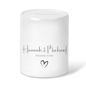 May include: White ceramic money box with a slot on top. The box has black text that reads "Hannah & Michael Wedding Fund" and a small black heart.