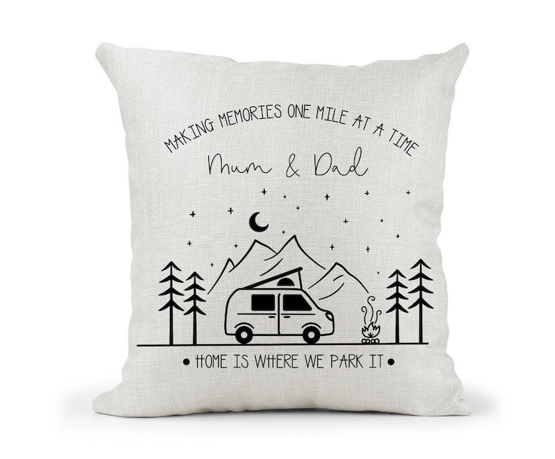 May include: White cushion with black text that reads "Making memories one mile at a time Mum & Dad" and a black illustration of a camper van, mountains, trees, and a campfire. The text at the bottom reads "Home is where we park it."