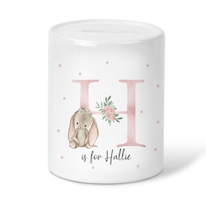 May include: White ceramic money box with a pink letter H decorated with pink flowers and a small brown bunny. The text "H is for Hallie" is printed below the letter.