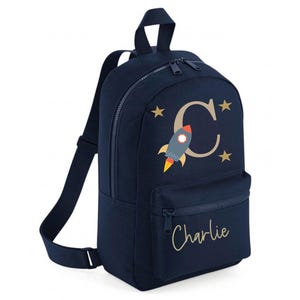 May include: A navy blue backpack featuring a rocket ship and the letter "C" design. The front pocket has the name "Charlie" in gold script. Gold stars are scattered around the rocket and letter.