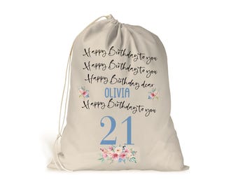 May include: A beige drawstring bag with the text "Happy Birthday to you" repeated four times, with the name "Olivia" in the third line. The number "21" is printed in blue with a floral design below it.