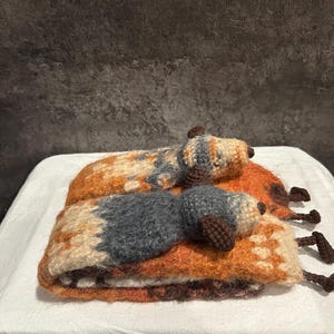 May include: Two handmade, crocheted dog-shaped pillows. The dogs are lying down, with brown, orange, and gray color patterns. The pillows are on a white surface, with a dark gray background.