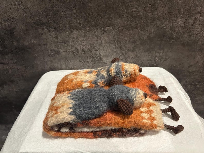May include: Two handmade, crocheted dog-shaped pillows. The dogs are lying down, with brown, orange, and gray color patterns. The pillows are on a white surface, with a dark gray background.