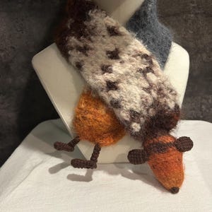 May include: A handmade scarf with a fox design. The scarf features a brown and white patterned section, a gray section, and an orange fox head with brown ears and legs. The scarf is displayed on a white mannequin.