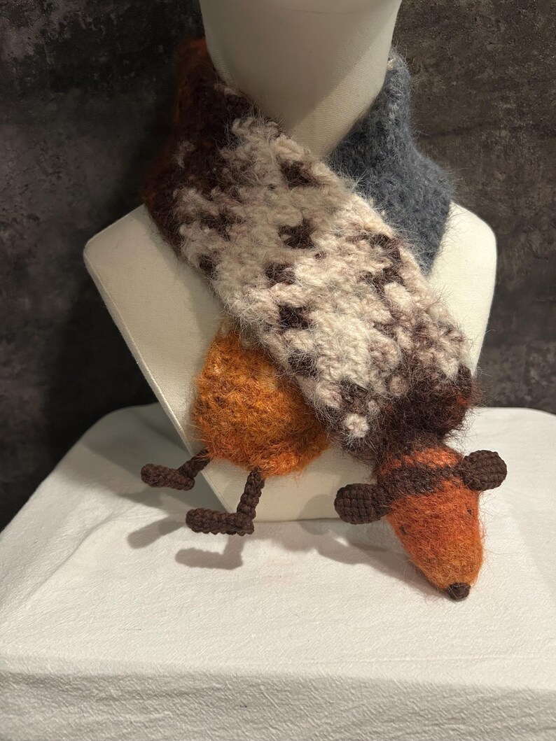 May include: A handmade scarf with a fox design. The scarf features a brown and white patterned section, a gray section, and an orange fox head with brown ears and legs. The scarf is displayed on a white mannequin.