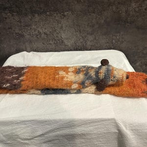 May include: A handmade, long, knitted fox-shaped pillow in shades of brown, orange, and cream. The fox has a small, round head with a dark brown nose and a small, round brown ball on top of its head. The pillow is lying on a white surface.