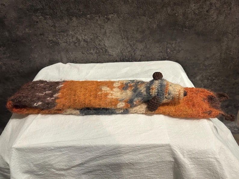 May include: A handmade, long, knitted fox-shaped pillow in shades of brown, orange, and cream. The fox has a small, round head with a dark brown nose and a small, round brown ball on top of its head. The pillow is lying on a white surface.