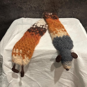 May include: Two handmade, knitted fox-shaped toys. One is a long, orange, brown, and cream-colored fox tail with brown legs. The other is a gray fox with brown ears and a cream-colored tail tip. The toys are on a white surface.