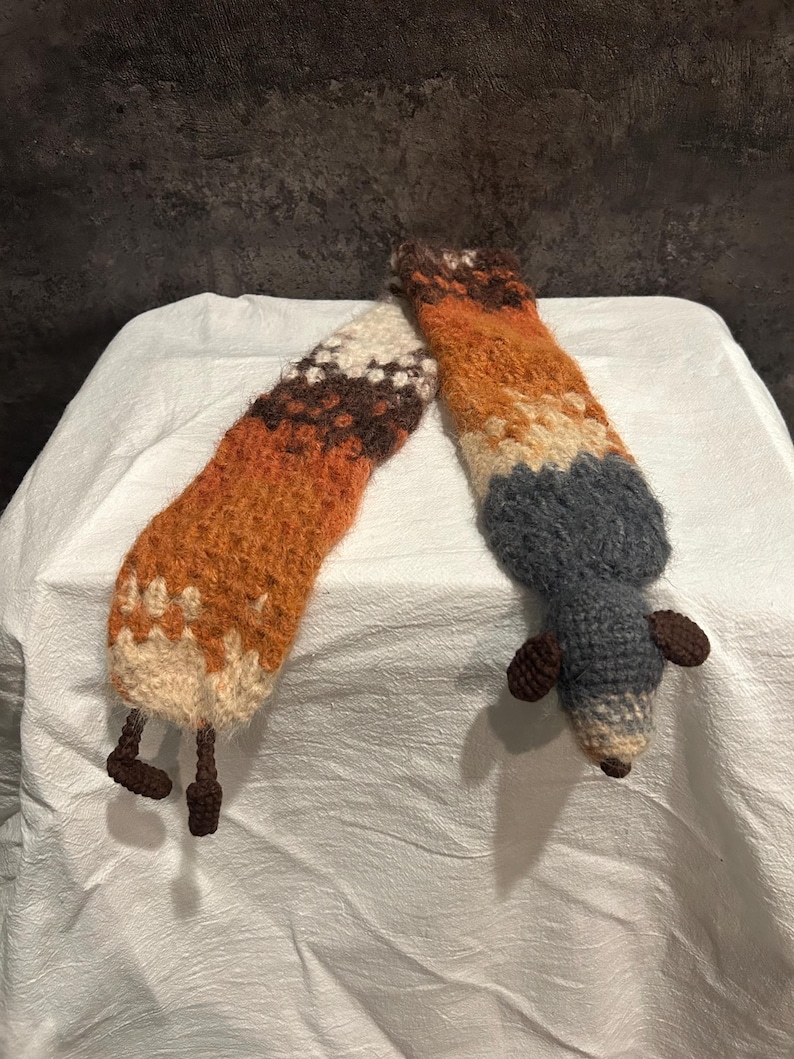 May include: Two handmade, knitted fox-shaped toys. One is a long, orange, brown, and cream-colored fox tail with brown legs. The other is a gray fox with brown ears and a cream-colored tail tip. The toys are on a white surface.
