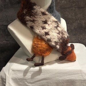 May include: A whimsical, handmade scarf with a unique design. The scarf features a combination of brown, white, and orange wool, with a pattern of dark brown shapes. It has a playful, animal-like design with orange legs and feet.