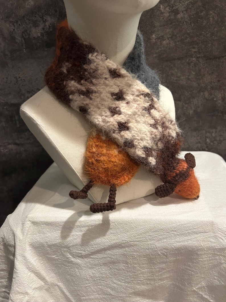 May include: A whimsical, handmade scarf with a unique design. The scarf features a combination of brown, white, and orange wool, with a pattern of dark brown shapes. It has a playful, animal-like design with orange legs and feet.