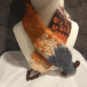 May include: A handmade scarf in the shape of a fox. The scarf features orange, tan, brown, and gray sections, with a fox tail design at the end. The scarf is displayed on a mannequin head.