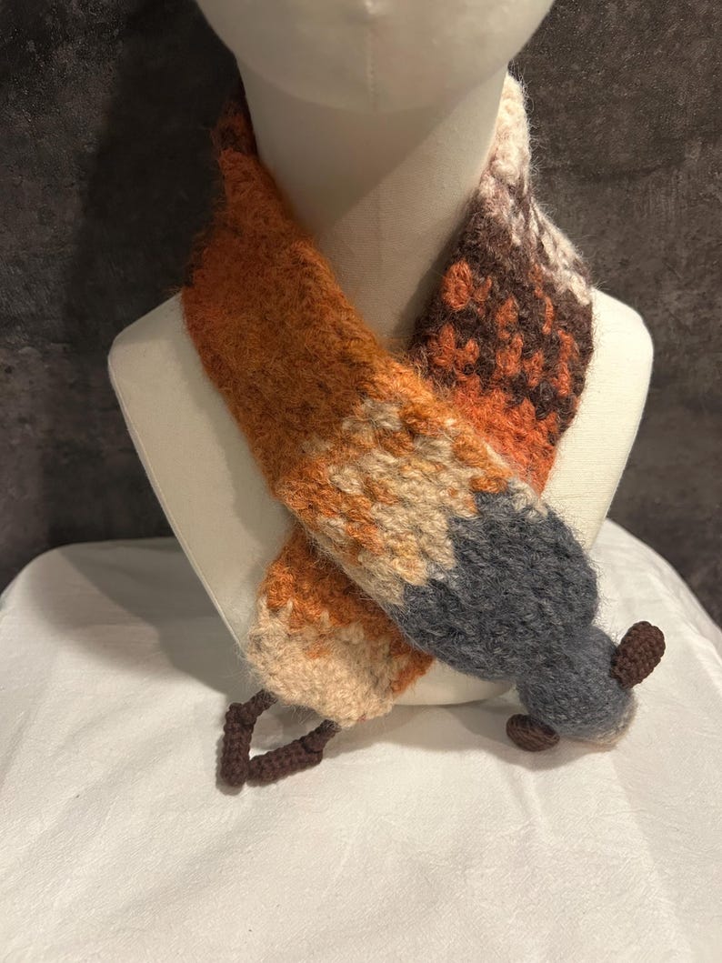 May include: A handmade scarf in the shape of a fox. The scarf features orange, tan, brown, and gray sections, with a fox tail design at the end. The scarf is displayed on a mannequin head.