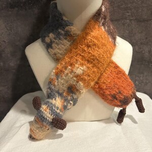 May include: A handmade fox scarf in shades of orange, brown, and blue, displayed on a mannequin. The scarf features a textured, knitted design with a fox head and tail details. The fox scarf is a unique accessory.