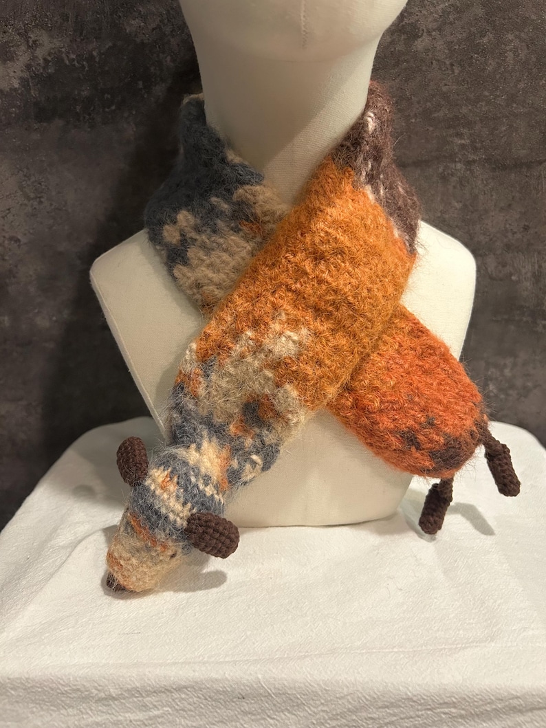 May include: A handmade fox scarf in shades of orange, brown, and blue, displayed on a mannequin. The scarf features a textured, knitted design with a fox head and tail details. The fox scarf is a unique accessory.
