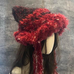 May include: A red and black fuzzy hat with a wide brim and long, dangling strands. The hat has a textured appearance with white accents. The hat is a unique accessory, perfect for adding a touch of flair to any outfit.