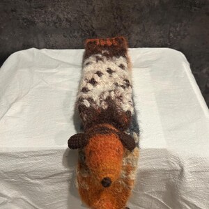 May include: A handmade, knitted hedgehog-shaped cozy in shades of orange, brown, white, and blue. The cozy has a long, rectangular shape with a hedgehog head and feet attached. The body is a mix of textured yarns, creating a unique, handcrafted look.