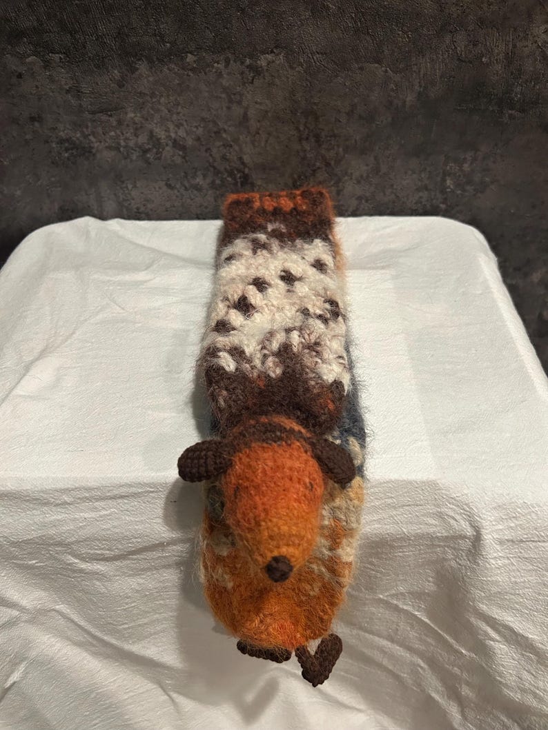 May include: A handmade, knitted hedgehog-shaped cozy in shades of orange, brown, white, and blue. The cozy has a long, rectangular shape with a hedgehog head and feet attached. The body is a mix of textured yarns, creating a unique, handcrafted look.