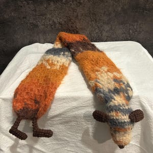 May include: A handmade, whimsical scarf in shades of orange, brown, and cream, with a fox-like design. The scarf features a striped pattern and a fox head and legs at each end, adding a playful touch to the accessory.