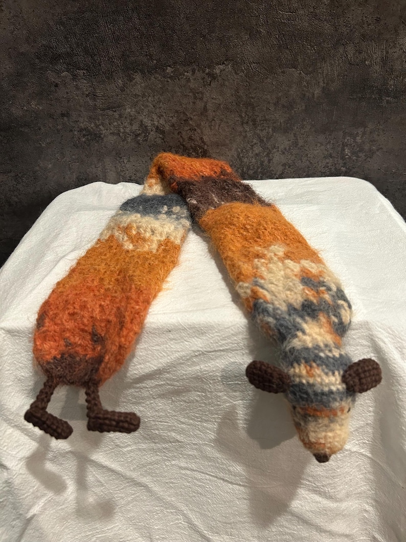 May include: A handmade, whimsical scarf in shades of orange, brown, and cream, with a fox-like design. The scarf features a striped pattern and a fox head and legs at each end, adding a playful touch to the accessory.