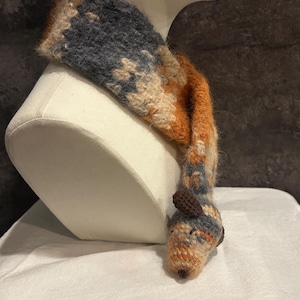 May include: A handmade scarf in shades of blue, orange, and beige, displayed on a mannequin head. The scarf features a whimsical animal head design at the end, adding a playful touch to the winter accessory.