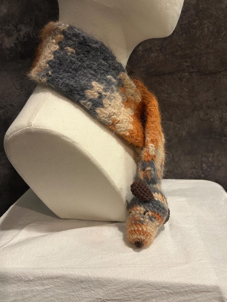 May include: A handmade scarf in shades of blue, orange, and beige, displayed on a mannequin head. The scarf features a whimsical animal head design at the end, adding a playful touch to the winter accessory.