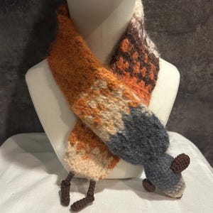 May include: A handmade scarf in the shape of a fox, displayed on a mannequin. The scarf features orange, brown, gray, and cream colors, with a textured, knitted appearance. The fox design includes a tail and small feet.