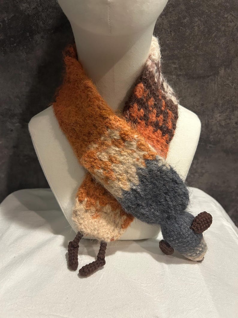 May include: A handmade scarf in the shape of a fox, displayed on a mannequin. The scarf features orange, brown, gray, and cream colors, with a textured, knitted appearance. The fox design includes a tail and small feet.