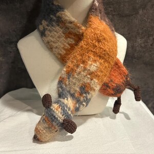 May include: A handmade scarf in the shape of a fox, displayed on a mannequin. The scarf features a color palette of orange, brown, beige, and blue. The fox design includes a head, ears, and tail.