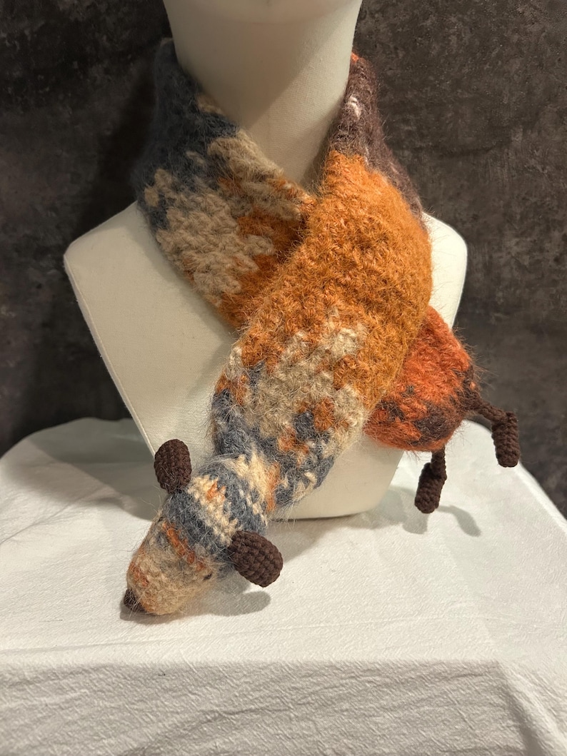 May include: A handmade scarf in the shape of a fox, displayed on a mannequin. The scarf features a color palette of orange, brown, beige, and blue. The fox design includes a head, ears, and tail.