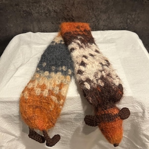 May include: A handmade, whimsical scarf in the shape of a fox. The scarf features a color gradient of orange, brown, cream, and blue. The fox's body and legs are crocheted, with the head and tail forming the ends of the scarf.