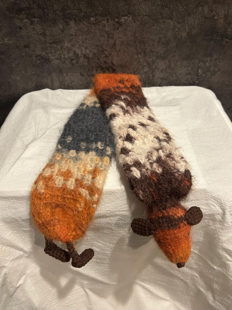 May include: A handmade, whimsical scarf in the shape of a fox. The scarf features a color gradient of orange, brown, cream, and blue. The fox's body and legs are crocheted, with the head and tail forming the ends of the scarf.