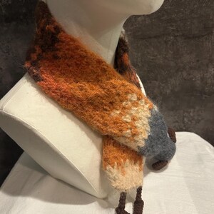 May include: A handmade fox scarf displayed on a mannequin. The scarf features a vibrant color palette of orange, brown, cream, and gray, mimicking a fox's fur. The scarf's design includes a pointed tail and fox-like features.