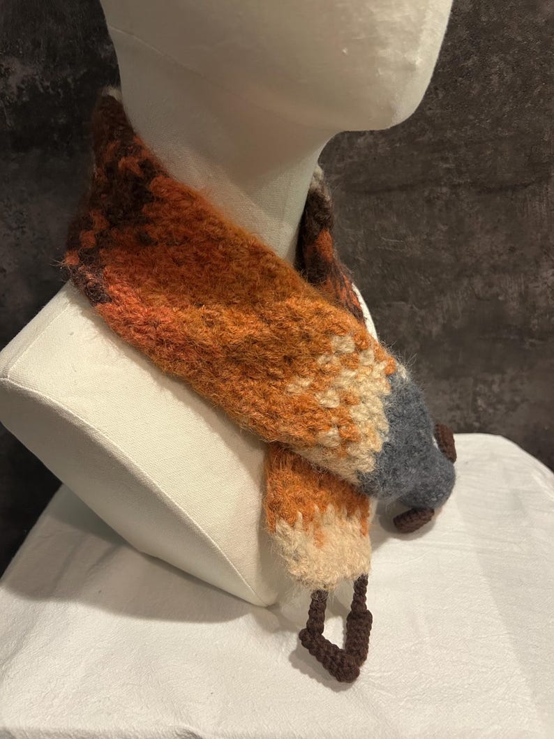 May include: A handmade fox scarf displayed on a mannequin. The scarf features a vibrant color palette of orange, brown, cream, and gray, mimicking a fox's fur. The scarf's design includes a pointed tail and fox-like features.
