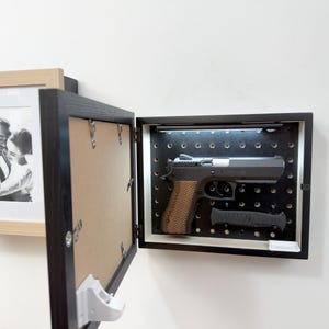 May include: A black wall-mounted storage box with a hinged picture frame door. The interior features a black pegboard back panel with integrated LED lighting, containing a semi-automatic pistol with a textured brown grip and a black folding knife.
