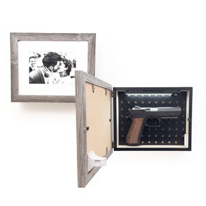 May include: A gray wooden picture frame with a black and white photo of a couple kissing. Next to it, a gray wooden gun safe with a black interior and a handgun with a brown grip.