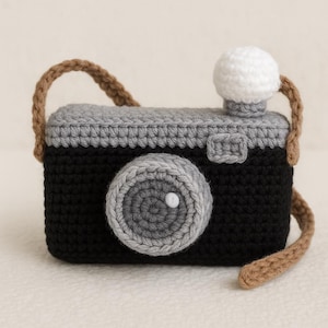 May include: A crocheted camera toy in black, grey, and white, with a brown strap. The camera has a large lens, a flash, and a detailed design. A charming handmade item, suitable as a child's toy or decorative piece.