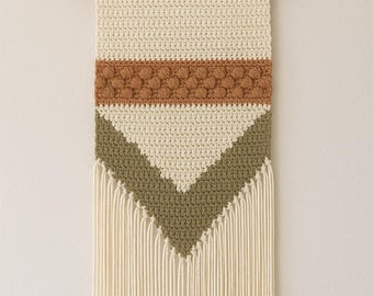 May include: A cream-colored, handmade macrame wall hanging with a wooden dowel and a natural fiber cord for hanging. The design features a brown textured stripe, a green chevron, and cream-colored fringe.